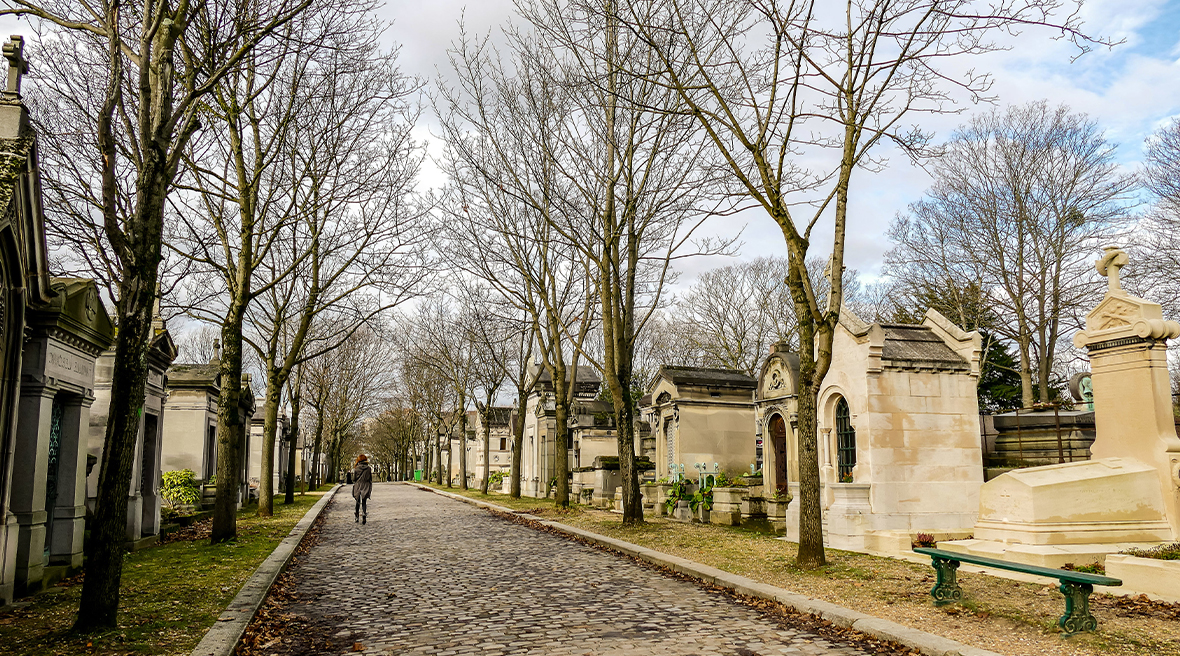Woman walking down a cobbled path lined with trees, tombs and gravestones on each side